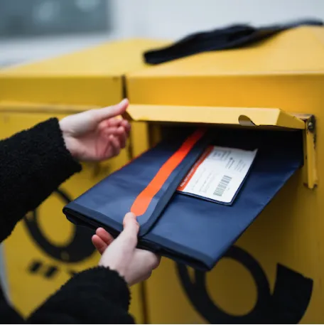 Eine Person wirft eine Boomerang Mehrwegversandtasche in einen gelben Briefkasten